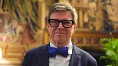 A close-up of Yann LeCun wearing thick rimmed black glasses, a navy suit with a dark blue bowtie standing in an opulent room at Buckingham Palace.