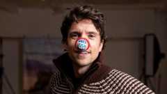 A close-up photo of Radio 1 DJ Greg James, he is wearing a brown and white stripped knitted jacket and has long curly brunette hair. He is staring directly at the camera and has a red nose on his nose that has a picture of a tandem bike on it and says Greg James.