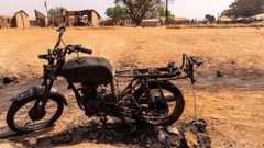 The charred frame of a burnt motorbike sits on dry, yellow grass near a small rural village.