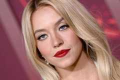 A close-up shot of Sydney Sweeney on a red carpet. She looks glamourous, with her blonde hair in a sleek, centre-parted style and a bold red lip.