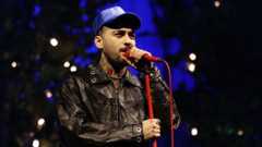 Zayn Malik wears a leather jacket and a blue hat. He is singing into a red microphone on a red microphone stand.