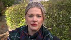 Siobhan Mackenzie looks straight ahead with a neutral expression. her blonde hair is tied back and she wears a dark green, blue and red tartan scarf around her neck. She is standing outside in front of a green hedge.