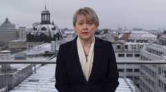 Foreign Secretary Yvette Cooper speaking from a conference in Munich. Shge wears a black overcoat with a white scarf and a blue beaded necklace. She has short, ash blonde hair with a fringe and two silver stud earrings. Snow-covered buildings can be seen behind her rooftop position. Copyright