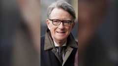 Peter Mandelson is wearing black glasses and a coat. He is pictured walking with a slight smile. His silver hair is being blown by the wind. The background is blurred.