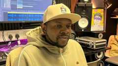 Xidus Pain in a recording studio. He is wearing a yellow hoodie with a yellow baseball cap and has a short neat black beard. Behind him is a mixing desk, speakers and a computer screen.