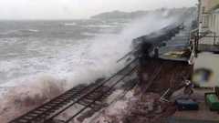 A damaged section of the coastal railway at Dawlish in 2014, with waves washing over broken track where the sea wall has collapsed. Houses sit close to the line on the right under a grey sky.