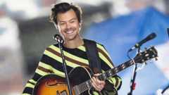 Harry Styles smiles in front of a microphone on a stand onstage as he holds a Gibson Les Paul guitar while wearing a striped green, black and brown top, with background bokeh behind, during a gig in New York in 2022.