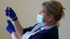 A stock picture of a female nurse in blue medical scrubs and white plastic apron. She is wearing blue gloves and a face mask and holding a vaccination needle in front of her.