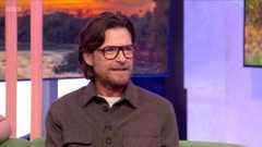 Jason Bateman has long hair swept back and wears glasses and a brown shirt