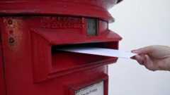 A hand posting a white envelope through a red post box opening.