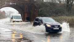 A car and a van drive through a deep puddle under a bridge, spraying water.