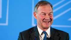 Rupert Lowe is wearing a suit blazer, a blue patterned tie and white shirt. He is smiling towards the right of the frame. There is a blue backing behind him.