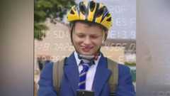 A boy in a blue blazer and striped tie has a yellow helmet on and a rucksack, and he is looking down at a phone. He is sticking his tongue out to one side of his mouth. He is outside.