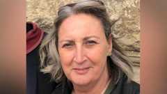 A woman with short grey hair wearing a black jacket, khaki green shirt, and a bag with a gold chain. She is smiling at the camera and has a pair of sunglasses on her head.