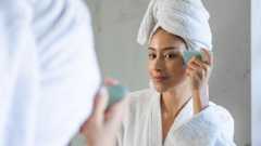 Woman wearing bathrobe and towel using a gua sha stone on her face, looking at herself in the mirror