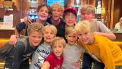A group of young boys in a small group smile at the camera. Many of them have long blonde hair and they are wearing T-shirt and shorts.