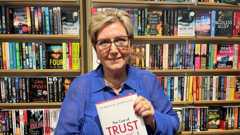 A woman with short blonde hair smiles at the camera while holding up her book, which says The Cost of Trust and Deborah Douglas on it. She wears a blue blouse and is sat in a book store in front of shelves of books.