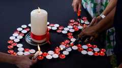 Small red and white candles are arranged in the shape of an Aids ribbon. A large white candle is in the centre. The candles are being lit by some disembodied hands.