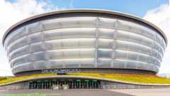 The Ovo Hydro is a tall, round silver building which looks like several large silver bands piled on top of one another. The entrance has several doorways and a sign above the entrance reads THE OVO HYDRO. The entrance has a small band of grass on top of it which snakes around the building. The land approaching the entrance is smooth grey pavement.