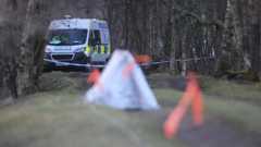 A police van is parked on a path in a wooded area. A small tent covering evidence is in the foreground.