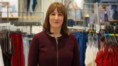 Chancellor Rachel Reeves stands in a clothes shop. She wears a burgundy jumper.
