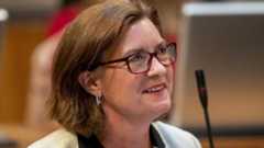 A head and shoulders picture of Eluned Morgan wearing red glasses and smiling in the Senedd chamber with a microphone in the background. She has a white jacket on, and the collars of a black shirt can be seen. She has silver earrings in.