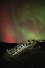 The Northern Lights in the north coast of Scotland. There are green and red stripes across the sky with the wooden frame of a boat in the foreground.