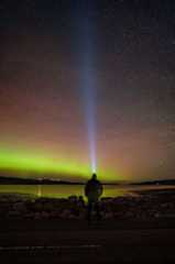 The Northern Lights in the north coast of Scotland. There are green and red hazes across the sky with Gary Macleod standing on a rocky beach in the foreground.