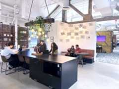 Modern office space with people working at a long desk with laptops, hanging plants and lights above. Cozy seating area nearby with drawings on the wall. Open layout featuring contemporary design elements like exposed brick and high ceilings.