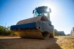 A construction roller on a dirt road at a construction site with buildings in the background and the sun shining behind.