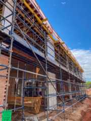 Construction site with scaffolding surrounding a building structure under construction, with exposed brick walls, concrete columns, wooden formwork, and a background of blue sky with some clouds.