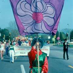 Person holding a purple flag with a flower design at a parade supporting LGBTQ+ community, with other parade participants and a banner in the background.