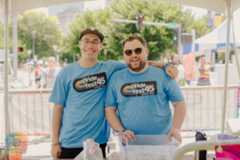 Two smiling men wearing blue Pride Fest 45 T-shirts, standing under a tent at an outdoor event, with trees and other people in the background.