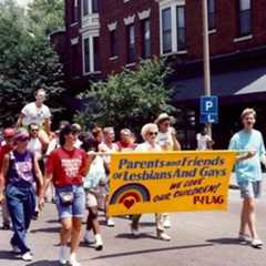 Group of people participating in a pride parade, carrying a banner that reads "Parents and Friends of Lesbians and Gays. We Love Our Children! PFLAG," with a rainbow heart and rainbow flag symbol.