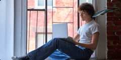 A young man casually sits on a window ledge with his laptop.