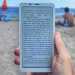 A photo of a hand holding a Boox Palma 2 e-reader with a beach scene with sunbathers visible in the background
