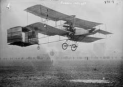 Photo shows a biplane flying low over the ground with a line of people watching in the background.