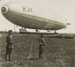 Two soldiers with rifles stand in front of a tethered dirigible.