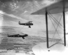 Photo shows a fuel line stretched between two airplanes in mid flight; one plane has a question mark painted on its side, and the other has the number one.