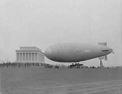 Photo shows a blimp tethered to the right of the Lincoln Memorial, with people around it.