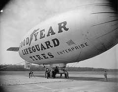 Photo shows a Goodyear blimp tethered to the ground while passengers climb into the gondola. Words on the blimp say "Lifeguard tires" and "Enterprise."