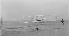 Photograph of the Wright Brothers biplane lifting off the ground with one brother lying in the plane and one standing next to the plane