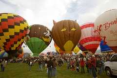 Color photo shows a crowd of people looking at five colorful balloons tethered on the ground.