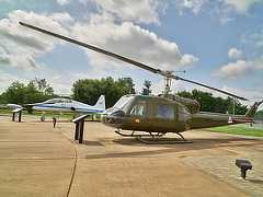 Color photo shows a helicopter (foreground) in foreground and a small airplane (background) parked on cement .