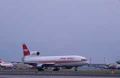 Color photo shows a side view of a large airplane, white with red stripe and tail fin, sitting on the airport runway