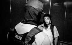 A man wearing a police vest looms over another man.