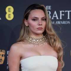 A woman with long, wavy hair wearing a glamorous pearl necklace and a strapless white gown poses confidently at the BAFTA Film Awards backdrop.