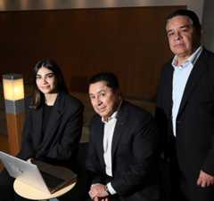 One woman and two men in a hallway, sitting on a chair with a laptop.