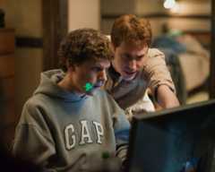 A man in a Gap sweatshirt with a dart in his teeth stares at a computer screen while another man leans in to look.