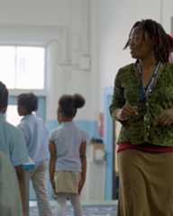 A woman with dreadlocks, wearing a green top and brown skirt, stands and gestures while several young children in school uniforms walk nearby in a brightly lit classroom.